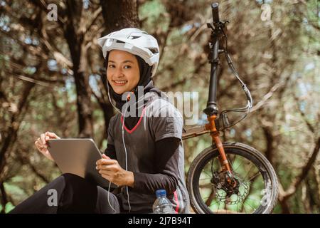 donna velata seduta a riposo in bicicletta indossando auricolari e tablet Foto Stock