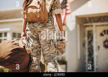 Vista posteriore di una mamma militare che tiene un orsacchiotto mentre si trova fuori casa con il suo bagaglio. Soldato patriottico femminile che torna a casa dopo serv Foto Stock