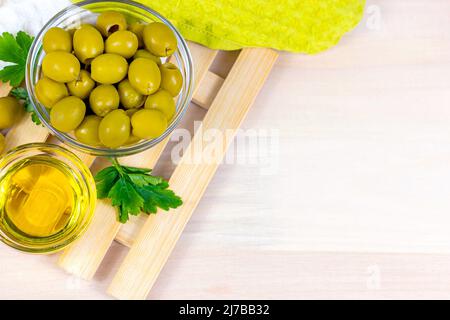 Molte olive verdi intere e olio d'oliva nelle ciotole di vetro su sfondo di legno chiaro in cucina vista dall'alto con spazio copia. Foto Stock