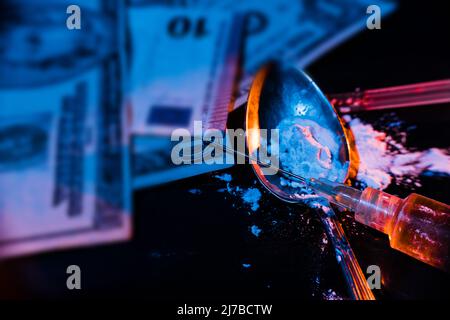 Siringa con Heroin cotto, polvere bianca in un cucchiaio e banconote su un tavolo scuro con illuminazione a due colori che suggerisce il proibito e illegale. Con Foto Stock