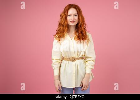 Foto di una bella giovane donna rossa con occhi blu che guarda la macchina fotografica vestita di tunica gialla in piedi su sfondo rosa. Foto Stock