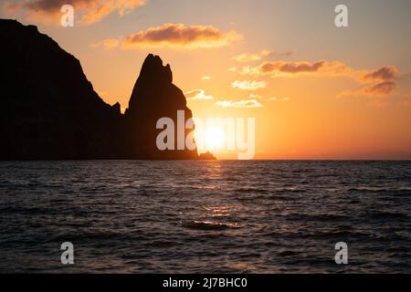 Un tramonto rosso acceso sul mare con scogliera vulcanica rocciosa. Astratta natura estate o primavera oceano mare sfondo. Piccole onde su acqua calda dorata Foto Stock