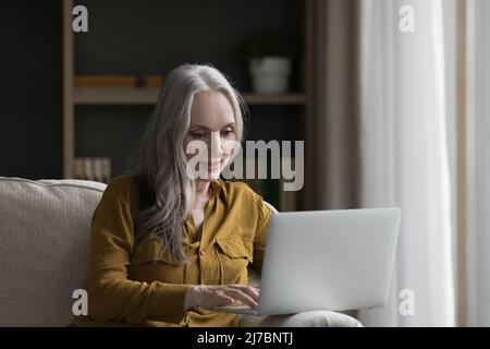 Donna dai capelli grigi focalizzata che usa l'app di teletrashy sul laptop Foto Stock