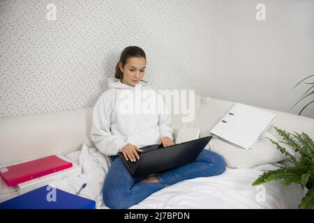 Una ragazza siede su un letto con liptopam e lavora, studia. Cartelle tutto intorno. Foto Stock