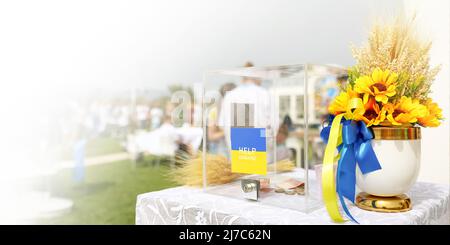 Beneficenza Ucraina raccolta fondi per donazioni in denaro in una scatola acrilica, vaso con fiori gialli e piante secche, nastro blu e giallo. Foto Stock