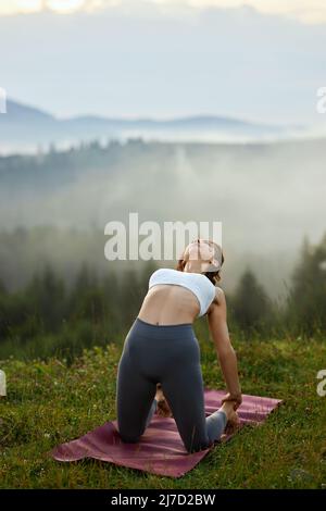 Vista frontale di una graziosa donna che indossa la parte superiore bianca e le leggings grigie che si piegano all'indietro toccando le gambe con le braccia all'aperto. Fit brunette ragazza tenuta cammello posa tra erba nella natura mattina. Concetto di yoga. Foto Stock