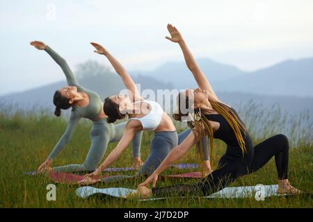 Vista laterale delle ragazze giovani che fanno la variazione crescente di guerriero posa nella natura del mattino. Le femmine si esercitano sul ginocchio raggiungendo le braccia indietro toccando le gambe posteriori ed estendendo le mani verso l'alto. Concetto di yoga. Foto Stock