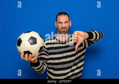 Bell'uomo latino che tiene il calcio con faccia arrabbiata, segno negativo che mostra dissimile con i pollici giù, concetto di rifiuto. Isolato su sfondo blu. Foto Stock