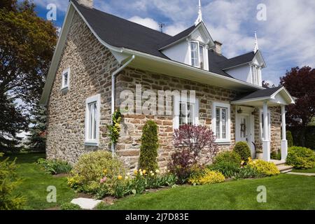 Old 1800s Canadiana cottage stile beige, marrone e marrone sfumato fieldstone casa con giardino paesaggistico anteriore in primavera. Foto Stock