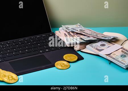 Computer portatile nero con alcuni bitcoins su di esso, un notebook con una bacchetta di dollari su un tavolo blu con uno sfondo verde e spazio copia. Formato orizzontale phot Foto Stock