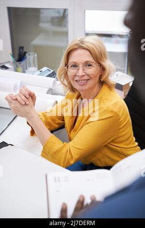 Donna d'affari matura in occhiali seduti alla sua scrivania e sorridente mentre ascoltava la relazione della sua collega Foto Stock