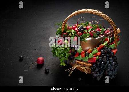 Cestino pieno di verdure, formaggio, uva e una bottiglia di vino su sfondo nero Foto Stock