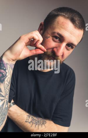 Ritratto verticale di un uomo tatuato di mezza età che tocca i baffi marroni e guarda la fotocamera su sfondo grigio. Foto di alta qualità Foto Stock