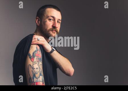 Gangster uomo sgucioso in una t-shirt nera che mostra ai tatuaggi della macchina fotografica sulla sua spalla su sfondo grigio scuro. Foto di alta qualità Foto Stock