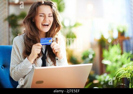 Green Home. Giovane donna felice con capelli ondulati lunghi con computer portatile e carta di credito seduta in una poltrona blu nel moderno soggiorno in giornata di sole. Foto Stock