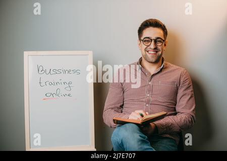 Insegnante con occhiali che tiene un libro in mano seduto a casa su una sedia accanto a una lavagna bianca che legge istruzione, apprendimento, studio. Parete bianca Foto Stock