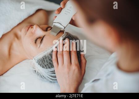 Vista dall'alto di una donna che riceve il peeling facciale con cavitazione a ultrasuoni nel salone termale. Faccia felice. Cosmetologia procedura di bellezza. Cura della pelle del viso. Foto Stock