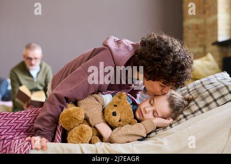 Giovane mamma attenta dando il suo adorabile figlio addormentato buona notte bacio mentre si piega su di lui contro senzatetto uomo maturo Foto Stock