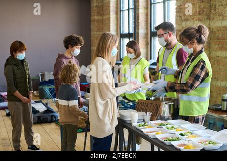 Gruppo di giovani volontari in maschere e guanti che spargono cibo cotto tra i rifugiati in piedi davanti a tavola con contenitori Foto Stock