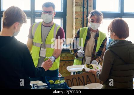 Volontario maschile in uniforme, maschera e guanti passando mela a ragazzo, mentre la giovane donna mettere cibo in contenitore di rifugiato femminile Foto Stock