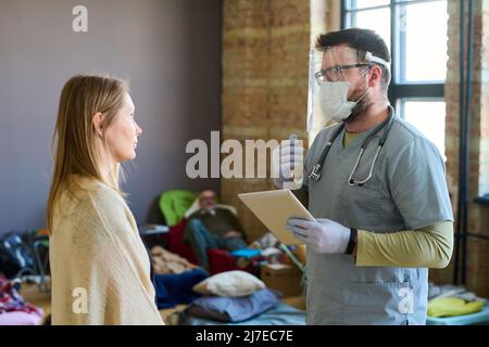 Medico di sesso maschile con tavoletta che dà consigli medici alla giovane rifugiata di sesso femminile malata nel campo e spiega come trattare la sua malattia Foto Stock