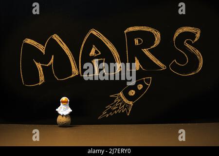 Una figura di astronauta meditante seduto su un pianeta. Sullo sfondo, una lavagna nera con scritta sfocata Benvenuto. Colonizzazione di Marte conc Foto Stock