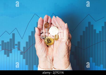 Le mani di femakes rugose tengono una manciata di bitcoins e monete d'oro. Business chart blu sullo sfondo. Il concetto di banca e di investimento. Foto Stock