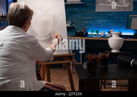 Insegnante d'arte pensionato concentrato che fa l'arte creativa nel disegno a matita ancora vita vase modello su tela di carta su cavalletto. Donna anziana che ombreggiano schizzo in studio di luce in classe sera sensazione di ispirazione. Foto Stock