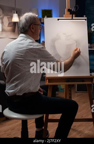 Uomo anziano che frequenta un laboratorio di arte serale che fa disegno originale del vaso usando matita hb su carta e cavalletto. Ritratto di artista anziano seduto su sgabello tracciare contorno di vaso in gesso. Foto Stock