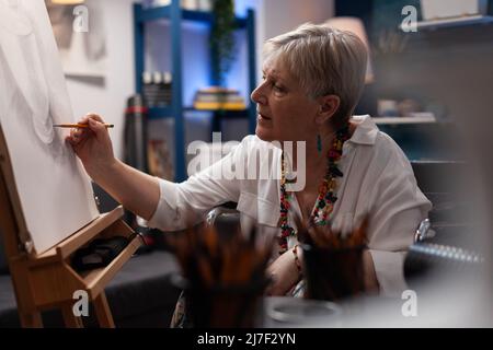 Artista anziano ombreggiando il modello di natura statica del vaso usando la matita su tela di carta in laboratorio creativo domestico. Donna anziana sensazione creativa disegno carbone schizzo su cavalletto in studio d'arte. Foto Stock