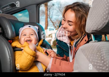 Ritratto di felice madre caucasica prepara il suo piccolo sorridente bambino carino per un viaggio in auto. Una donna allaccia le cinture di sicurezza in un seggiolino per bambini. Visualizza insi Foto Stock