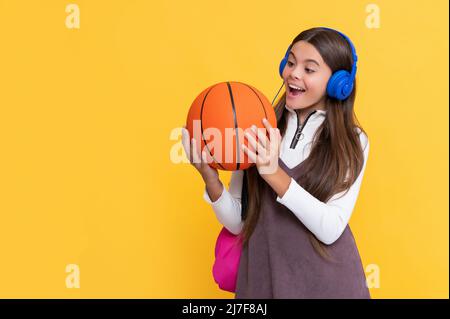 il bambino ha scioccato in cuffie con zaino scuola e palla da basket su sfondo giallo Foto Stock