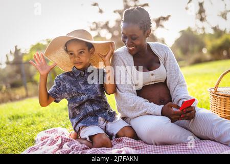 Felice madre incinta africana che passa il tempo con suo figlio fare un picnic durante il fine settimana nel parco pubblico - Maternity e genitori stile di vita concetto Foto Stock