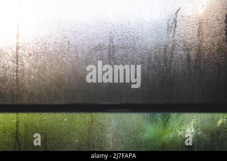 gocce d'acqua di condensa di umidità sul vetro del finestrino viste con la tendina avvolgibile e bokeh sul cortile nello scatto di sfondo a deth poco profondo di campo Foto Stock