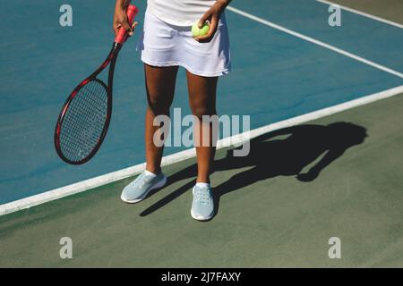 Sezione bassa di giovane donna afroamericana in piedi con racchetta da tennis e palla a campo Foto Stock