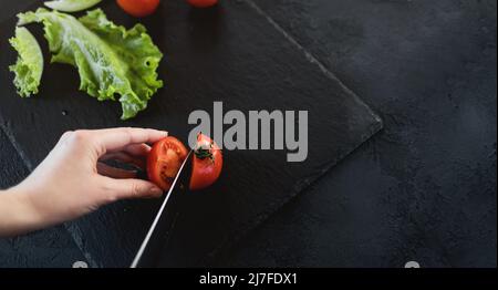 La donna sta tagliando i pomodori ciliegini per l'insalata sul bordo nero dell'ardesia. Cucina vegana Foto Stock