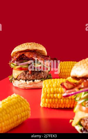 Cheeseburger fresco con lattuga, cetriolo, pomodoro, cipolla e pancetta su sfondo rosso vibrante. Guarnito con mais sulla pannocchia Foto Stock