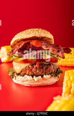 Cheeseburger fresco con lattuga, cetriolo, pomodoro, cipolla e pancetta su sfondo rosso vibrante. Guarnito con mais sulla pannocchia Foto Stock