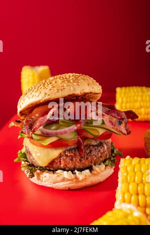 Cheeseburger fresco con lattuga, cetriolo, pomodoro, cipolla e pancetta su sfondo rosso vibrante. Guarnito con mais sulla pannocchia Foto Stock