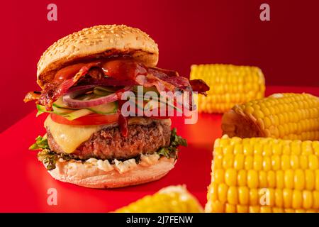 Cheeseburger fresco con lattuga, cetriolo, pomodoro, cipolla e pancetta su sfondo rosso vibrante. Guarnito con mais sulla pannocchia Foto Stock