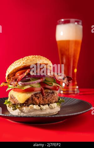 Cheeseburger fresco con lattuga, cetriolo, pomodoro, cipolla e pancetta su sfondo rosso vibrante. Abbinata alla birra Foto Stock