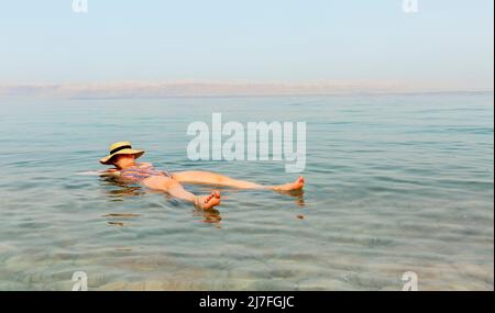 Donna bionda con cappello che galleggia nelle acque turchesi del Mar Morto Foto Stock