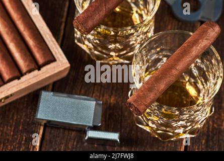 Vista di una scatola di legno con sigari cubani, un accendisigari e una taglierina. Ancora vita con due bicchieri di whiskey sul tavolo. Foto Stock