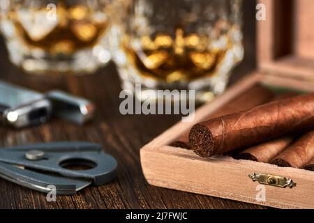 Vista di una scatola di legno con sigari cubani, un accendisigari e una taglierina. Ancora vita con due bicchieri di whiskey sul tavolo. Foto Stock