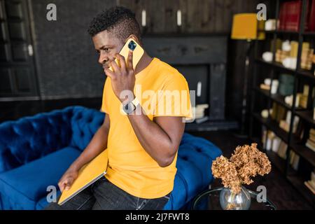 Sorridente giovane studente afroamericano o uomo d'affari che fa professionale chiamata di lavoro sul portatile giallo a casa godendo aziendale conversazione Foto Stock