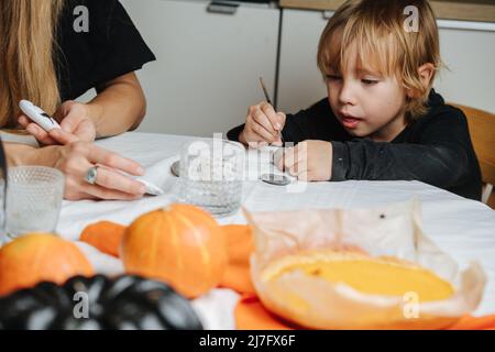 Piccolo figlio seduto dietro un tavolo rotondo con la mamma, preparando decorazioni per Halloween. Pittura su pietre, zucche e bottiglie. Foto Stock