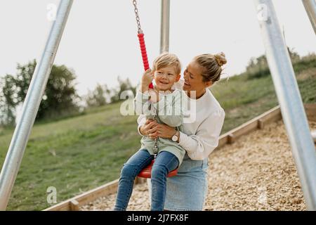 Felice madre che dondola figlio al parco giochi Foto Stock