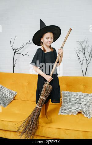 Ragazza piccola carina vestita come una strega che tiene una scopa, in posa  per una foto. Il colore dei calzini che si abbina al colore del divano su  cui sta in piedi Foto stock - Alamy