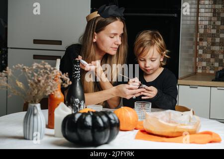 Mamma e suo figlio seduto dietro un tavolo rotondo, preparando decorazioni per Halloween. Pittura su pietre, zucche e bottiglie. Foto Stock