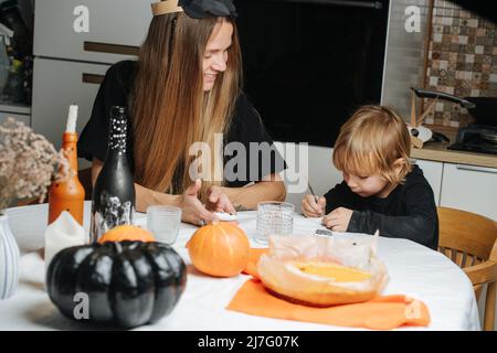 Mamma e suo figlio seduto dietro un tavolo rotondo, preparando decorazioni per Halloween. Pittura su pietre, zucche e bottiglie. Foto Stock